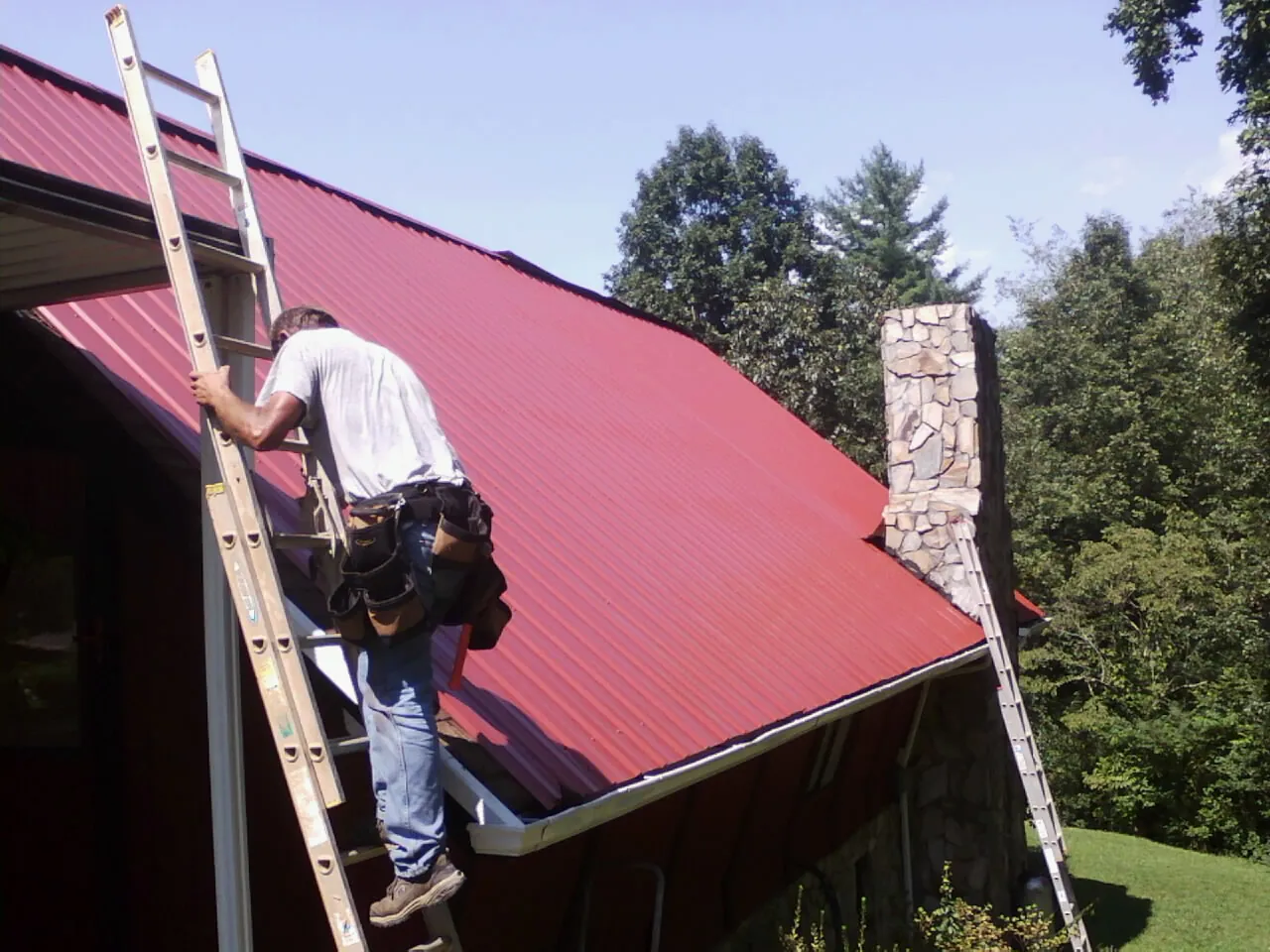 Roofer on a ladder beside a red metal roof and stone chimney — Soffit & Fascia Repair by Roofing Brothers in Arimo
